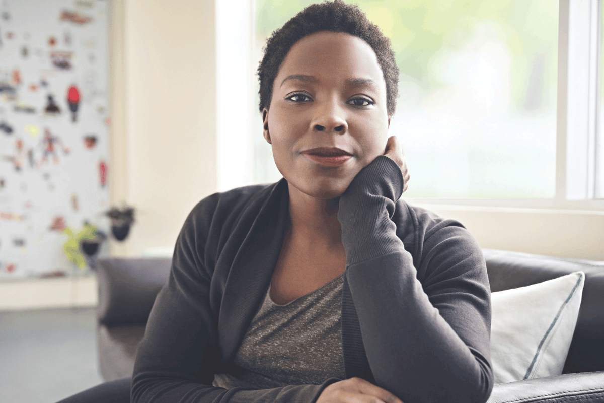 Assise sur un canapé, une femme noire pose un regard assuré sur l'objectif, un léger sourire aux lèvres, la tête appuyée sur