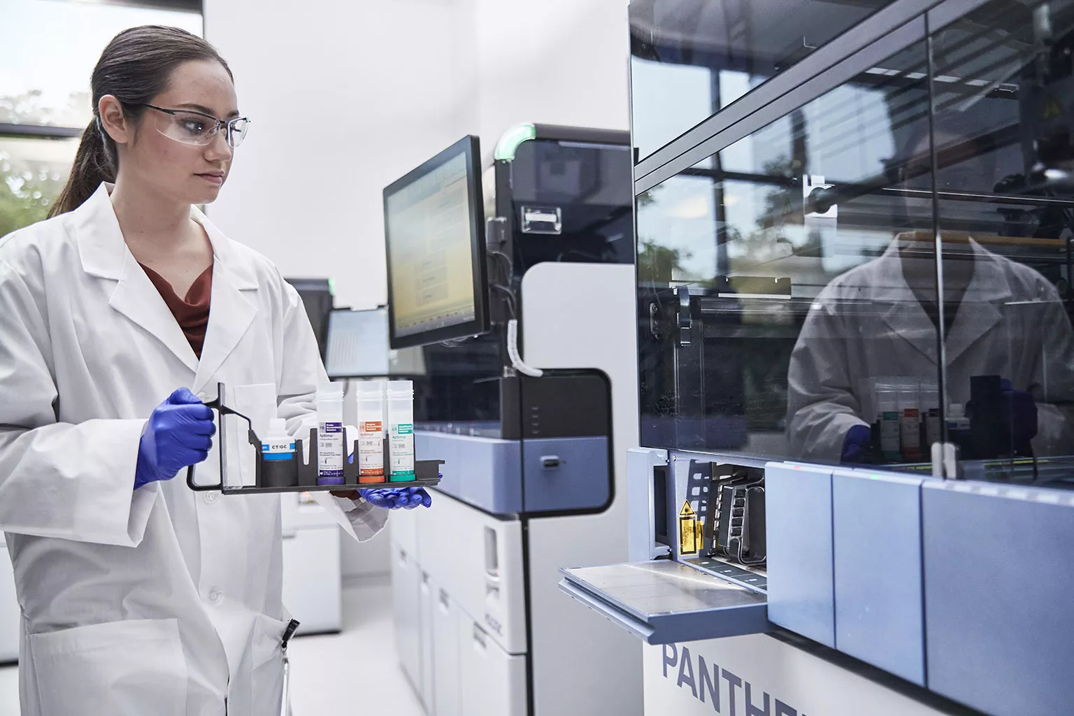 Une femme médecin tient un plateau de tubes à côté d’un système Panther dans un laboratoire.